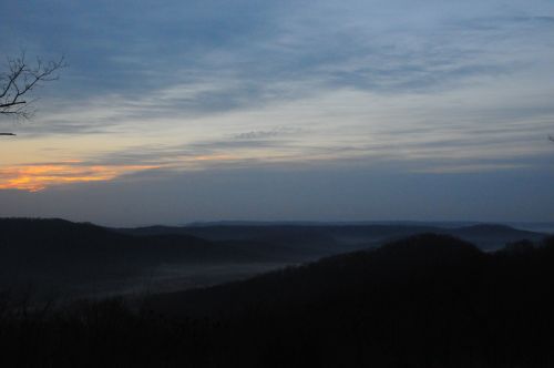 A serene landscape at dusk, featuring rolling hills shrouded in mist under a cloudy sky with hints of orange light.