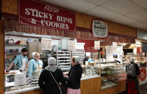 A bakery counter with staff serving sticky buns and cookies to customers in a cozy, rustic setting.