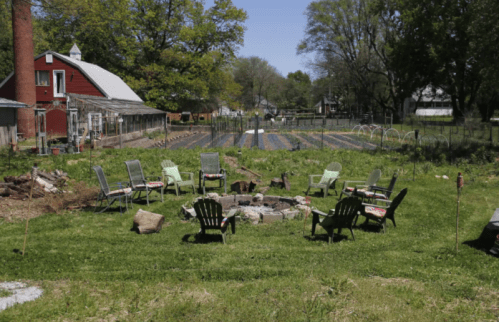 A sunny garden with a fire pit surrounded by chairs, overlooking rows of crops and a red barn in the background.