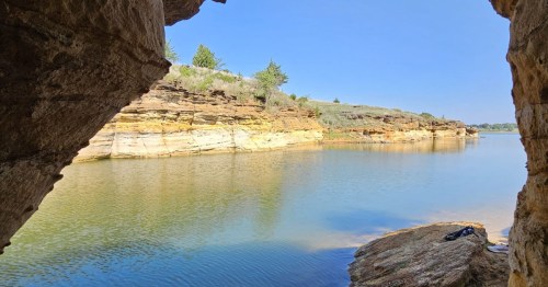 View of a calm lake and layered sandstone cliffs framed from inside a dark rocky cave opening.