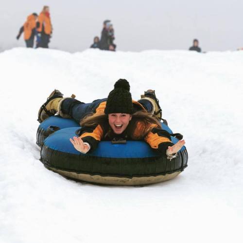 A person in winter attire joyfully sleds down a snowy hill on a blue inflatable tube.