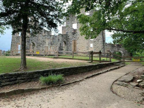 Ruins of a stone building surrounded by trees, with a pathway leading to the entrance and warning signs nearby.