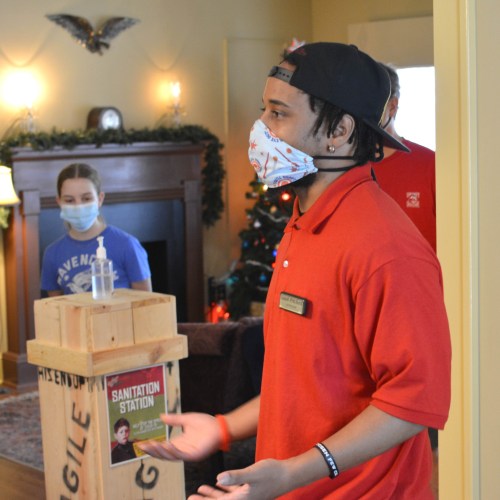 A young man in a red shirt and mask stands near a sanitation station, while another person in a mask is visible in the background.