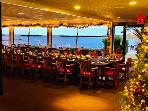 A festive dining room with a long table set for a meal, decorated with lights and a view of the water outside.