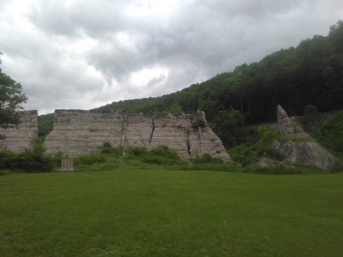 A grassy area with large, weathered rock formations and a backdrop of green hills under a cloudy sky.