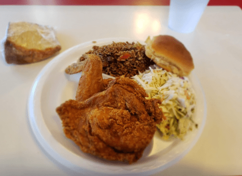 A plate with fried chicken, coleslaw, rice, a roll, and a slice of cake on a white table.