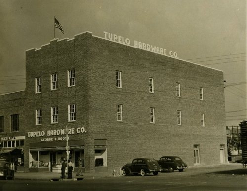 Historic black-and-white photo of the Tupelo Hardware Co. building, featuring a flag and vintage cars parked outside.