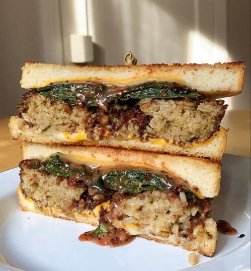 A stacked sandwich with crispy bread, filled with a savory mixture, cheese, and greens, drizzled with sauce.
