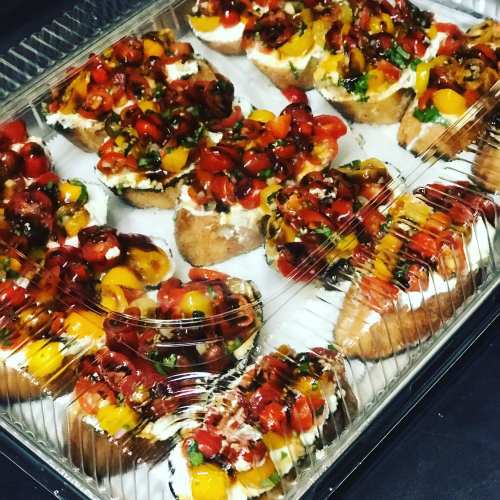 A tray of colorful bruschetta topped with tomatoes, herbs, and cheese, arranged neatly in a clear plastic container.