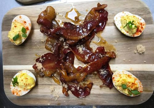 A wooden platter with crispy bacon strips drizzled in syrup, accompanied by deviled eggs topped with green onions.