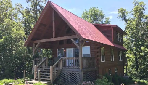 A wooden cabin with a red metal roof, surrounded by trees and greenery, featuring a spacious front porch.