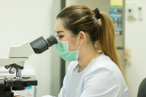 A woman in a mask examines a sample through a microscope in a laboratory setting.