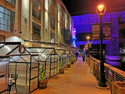 A lit riverside walkway with glass enclosures, festive decorations, and a brewery sign under a colorful night sky.