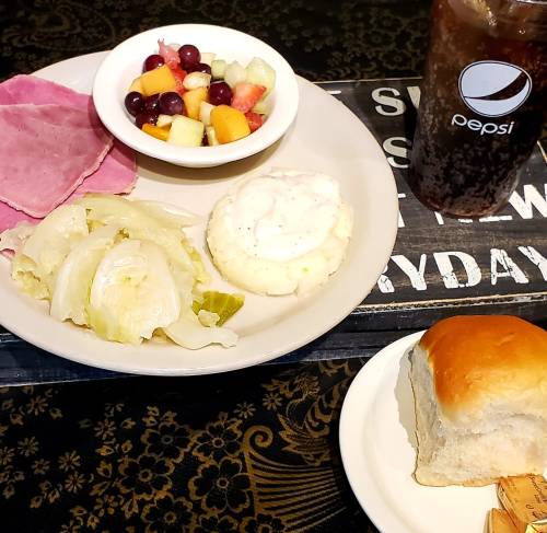A plate with sliced ham, cabbage, mashed potatoes with gravy, fruit salad, a roll, and a glass of Pepsi.