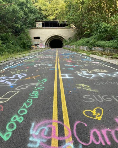 A colorful, graffiti-covered road leads to a dark tunnel, surrounded by lush greenery.