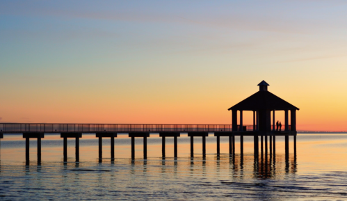 Silhouette of a pier with a gazebo at sunset, reflecting on calm water, with two people standing at the end.