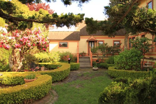 A charming garden with neatly trimmed hedges, colorful flowers, and a cozy house in the background under a clear sky.