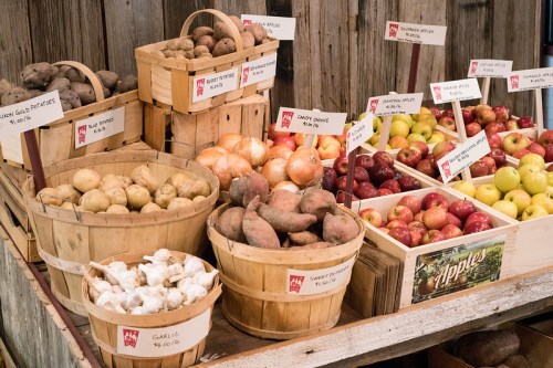 Baskets of various potatoes, garlic, and apples displayed on a rustic wooden table with price tags.