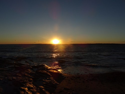Sunset over the ocean, casting golden light on the water and rocky shoreline.