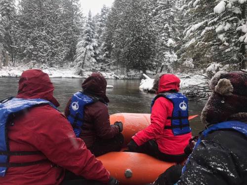 Group of people in life jackets sitting in a raft on a snowy river, surrounded by trees.