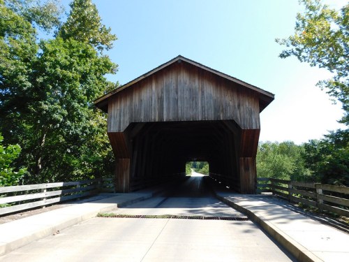 A wooden covered bridge with a clear blue sky and trees on either side, leading to an open road.