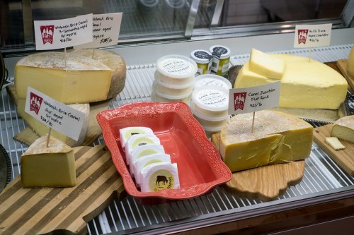 A display of various cheeses with price tags, including blocks and rounds, on wooden boards and a red dish.