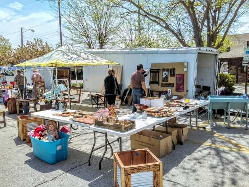 A bustling outdoor market with tables of handmade items, a blue storage bin, and people browsing under a yellow canopy.
