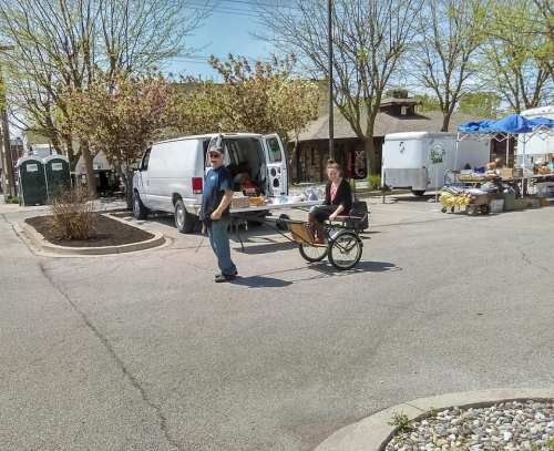 A person pushes a cart while another stands beside a van in a busy outdoor market area with trees in the background.