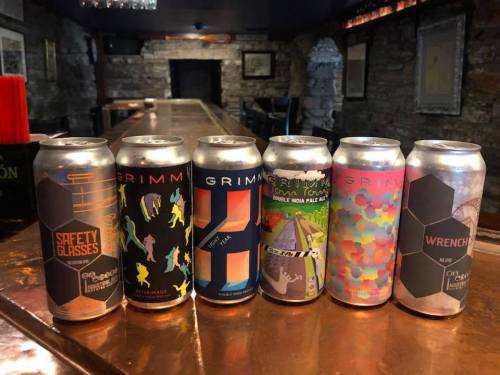 Six colorful beer cans lined up on a bar, featuring various designs and labels from Grimm Artisanal Ales.