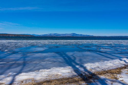 A serene winter landscape featuring a frozen lake, blue sky, and distant mountains under bright sunlight.