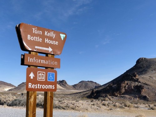 Signpost directing to Tom Kelly Bottle House, information, and restrooms, with mountains in the background.