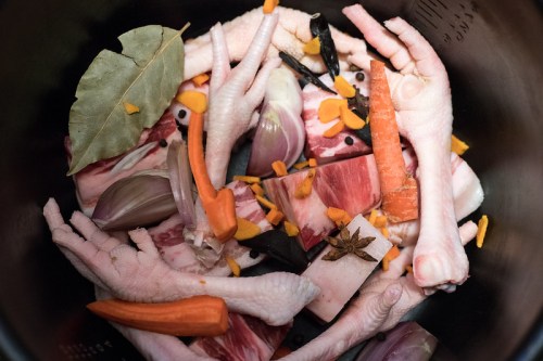 A close-up of raw meat, chicken feet, and vegetables, including carrots and onions, in a cooking pot.