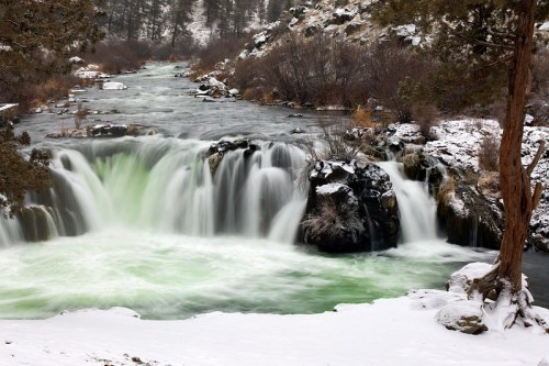 A serene waterfall cascades over rocks into a river, surrounded by snow-covered banks and trees in a winter landscape.