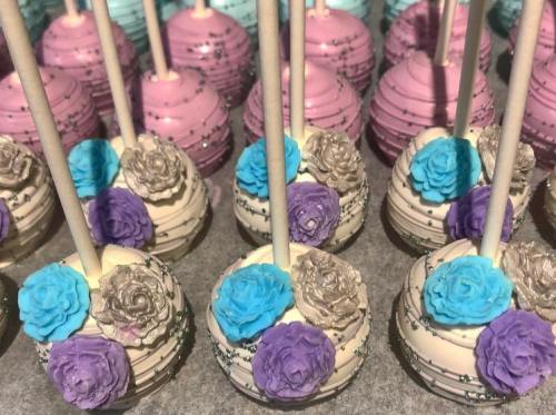 Colorful cake pops decorated with edible flowers in shades of blue, purple, and gold, arranged in neat rows.