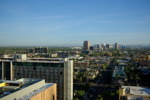 A panoramic view of a city skyline with modern buildings, streets, and mountains in the background under a clear blue sky.