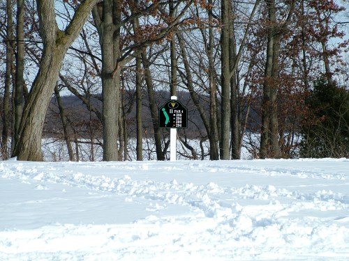 A snowy landscape with a golf course sign indicating hole 11, surrounded by bare trees and a distant view of hills.
