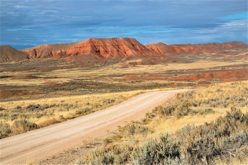 A winding dirt road leads through a vast landscape of red rock formations and dry grass under a blue sky.