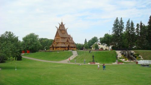 A wooden structure resembling a traditional Scandinavian church, surrounded by green grass and trees, with people walking nearby.