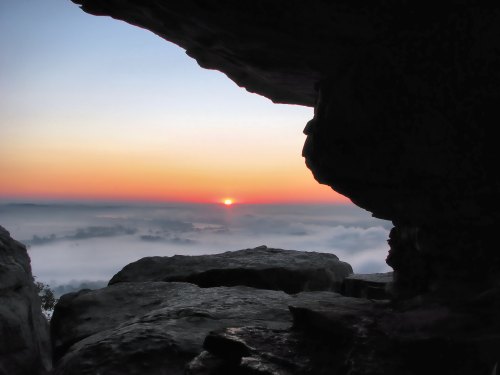 A serene sunset viewed through a rocky cave, with soft colors blending in the sky and misty landscape below.