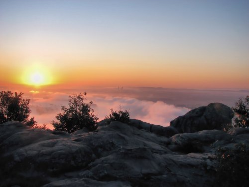 A stunning sunrise over a foggy landscape, with rocky terrain in the foreground and soft clouds below.