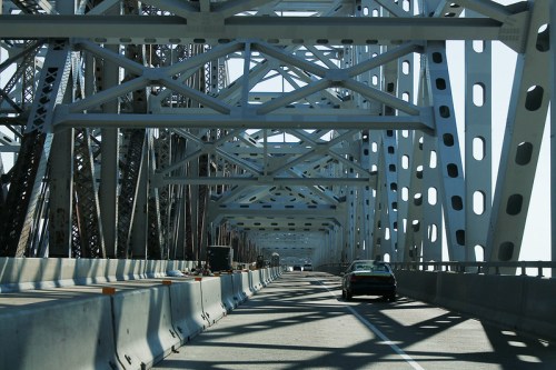 View of a bridge's metal framework with a car driving on the road, surrounded by construction barriers.