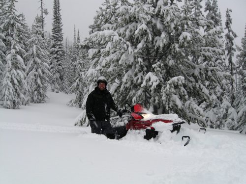 A person in winter gear stands next to a snowmobile in a snowy forest, surrounded by tall, snow-covered trees.