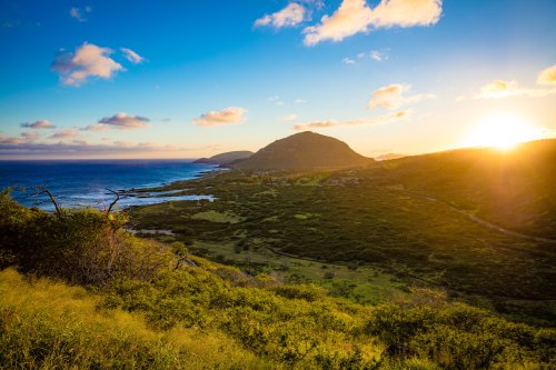 A scenic view of a coastal landscape at sunset, featuring mountains, ocean, and lush greenery under a colorful sky.