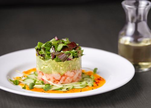 A layered dish featuring shrimp, avocado, and greens, drizzled with sauce, served on a white plate with a glass bottle.