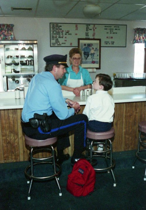 A police officer talks to a young boy at a diner counter, while a woman in an apron serves them.