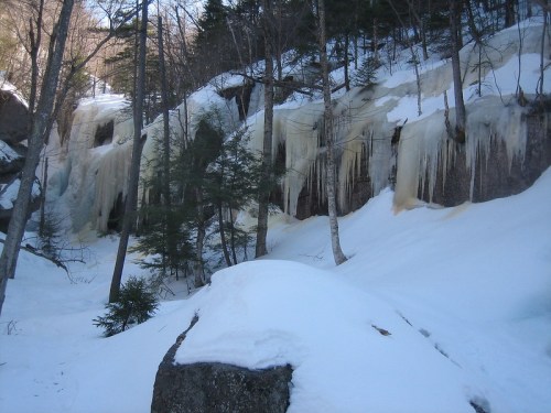 Icicles hang from a rocky cliff surrounded by snow-covered ground and trees in a winter landscape.