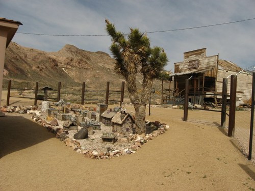 A desert scene featuring a miniature village, a Joshua tree, and an old wooden building in the background.