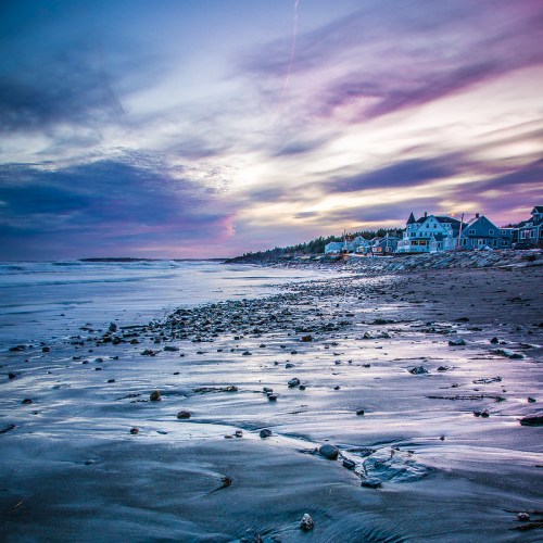 A serene beach at sunset, with colorful clouds and a rocky shore lined by charming houses.