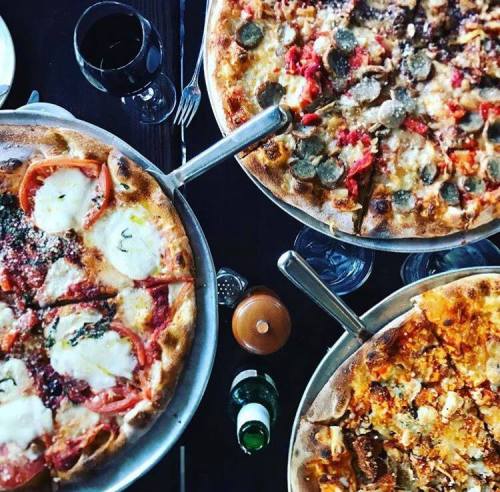 Three pizzas on metal trays, topped with various ingredients, alongside glasses of red wine and a bottle of beer.