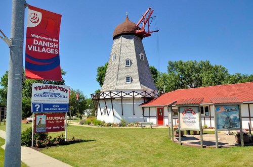 A Danish windmill stands in a village, surrounded by green grass and a welcome sign, under a clear blue sky.